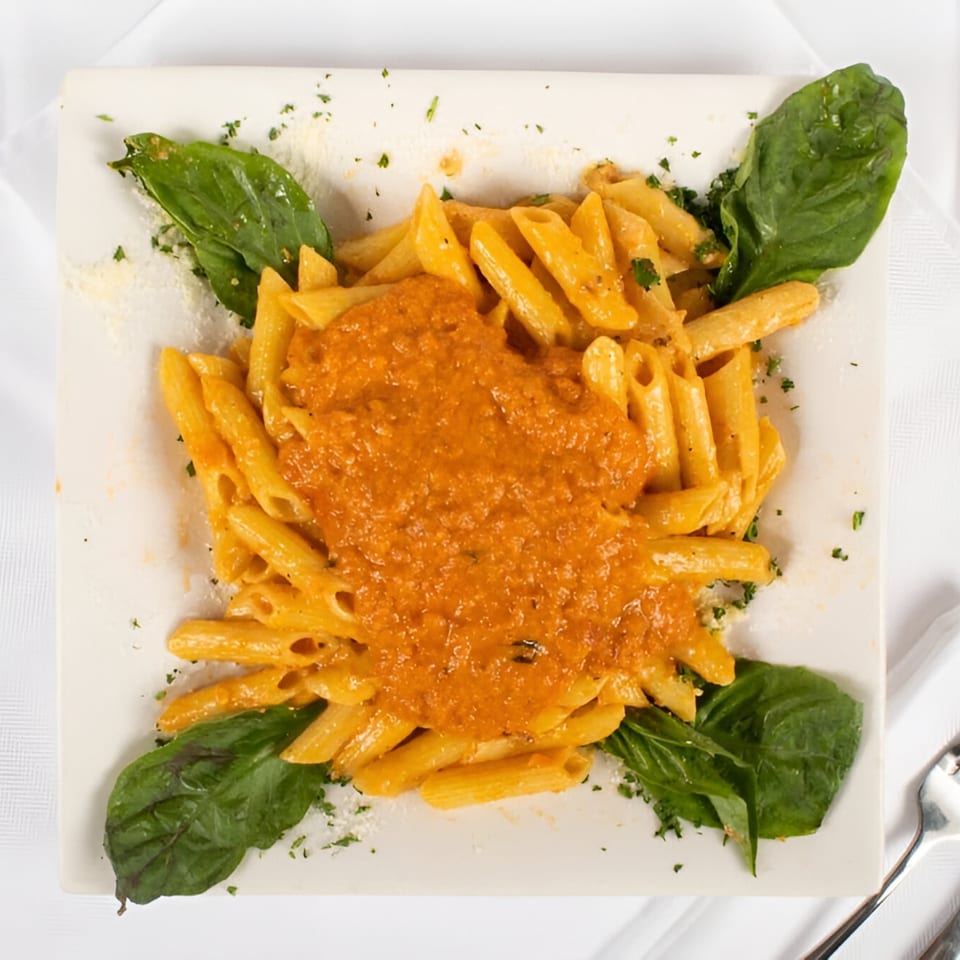 Penne pasta with orange sauce, garnished with fresh basil leaves and parsley on a white plate.